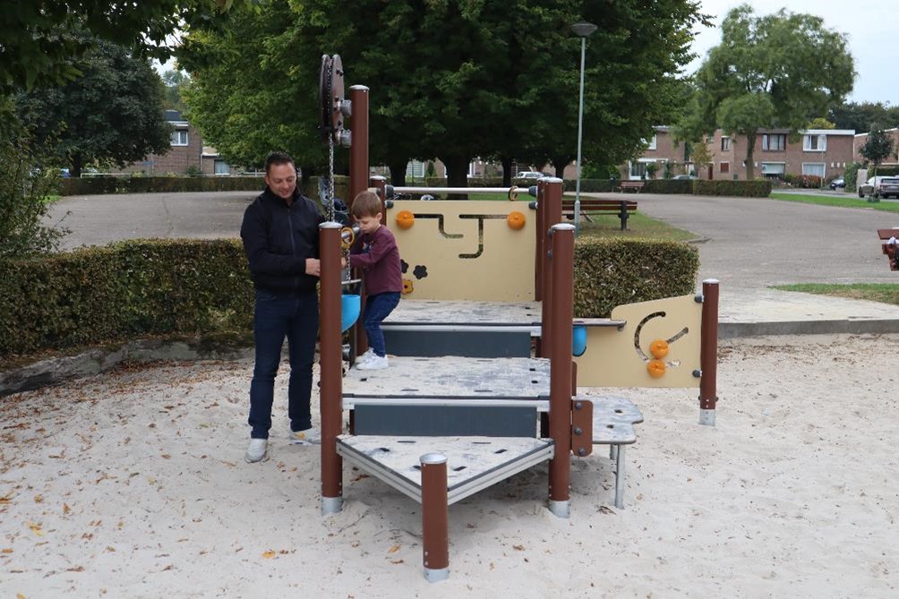 Speelplek met zandige bodem waarop speeltoestel staat met kind en man die samen een emmertje met zand aan ketting ophoog trekken